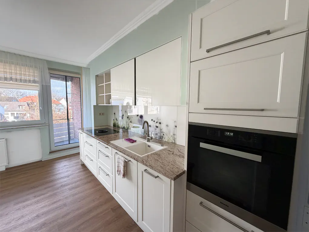 Bright kitchen with white cabinets, granite countertops, and a floral backsplash. A window and balcony door let in natural light.