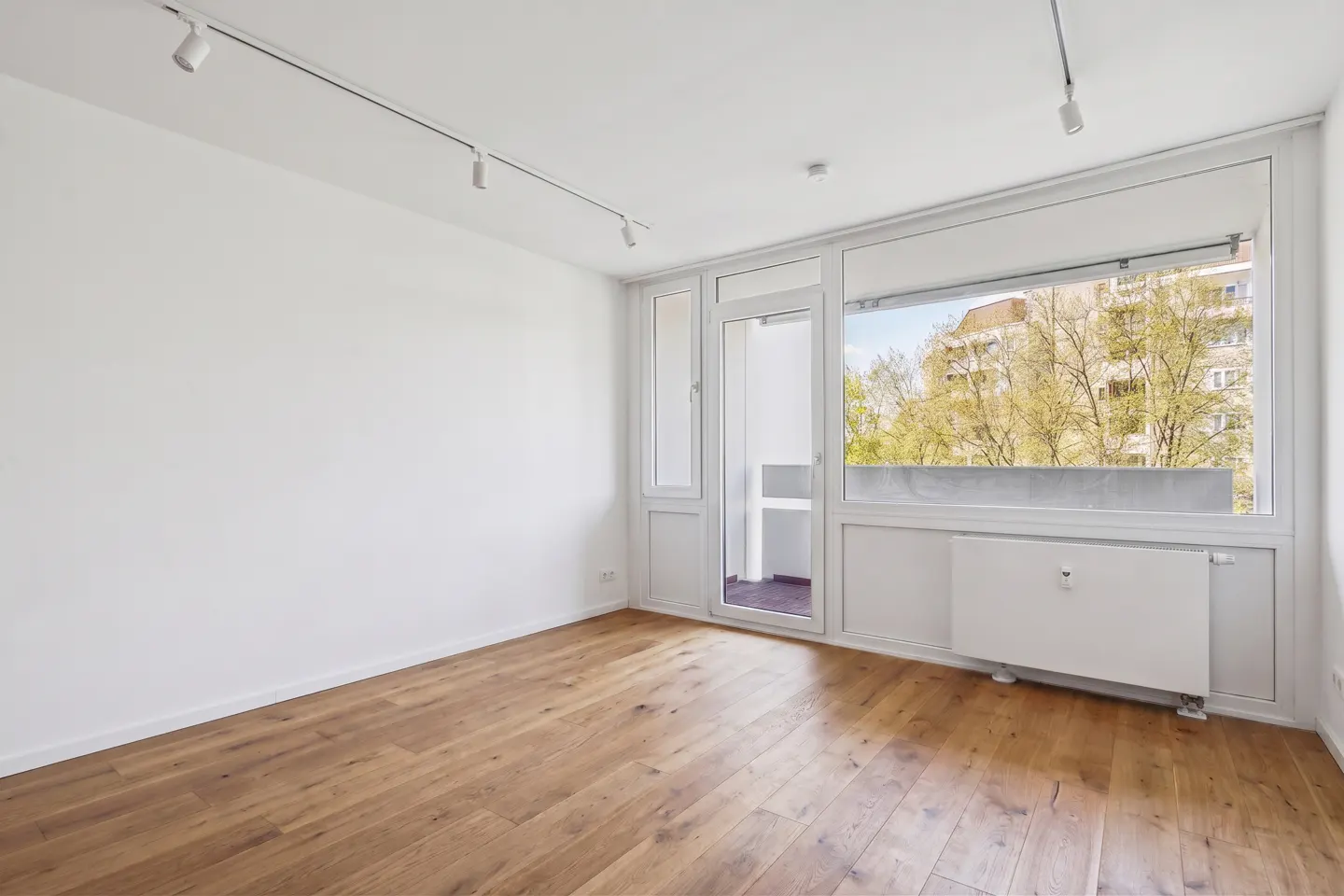 Bright, empty room with hardwood floors, white walls, and track lighting. A large window and door lead to a balcony with a view of trees and a building.