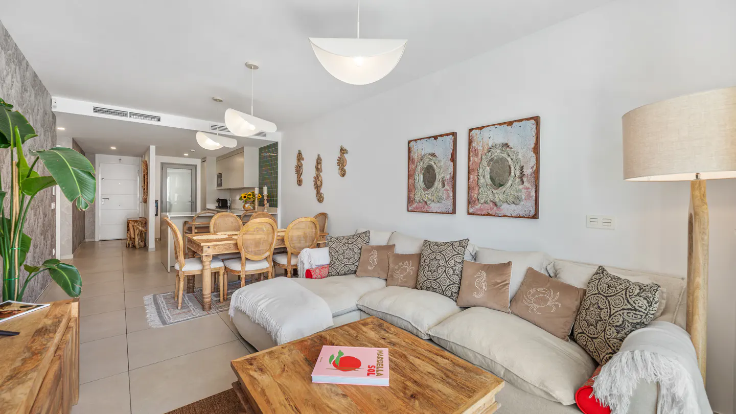 Bright living room with white walls, a beige sectional sofa with decorative pillows, and a wooden coffee table. A dining table and kitchen are visible in the background.