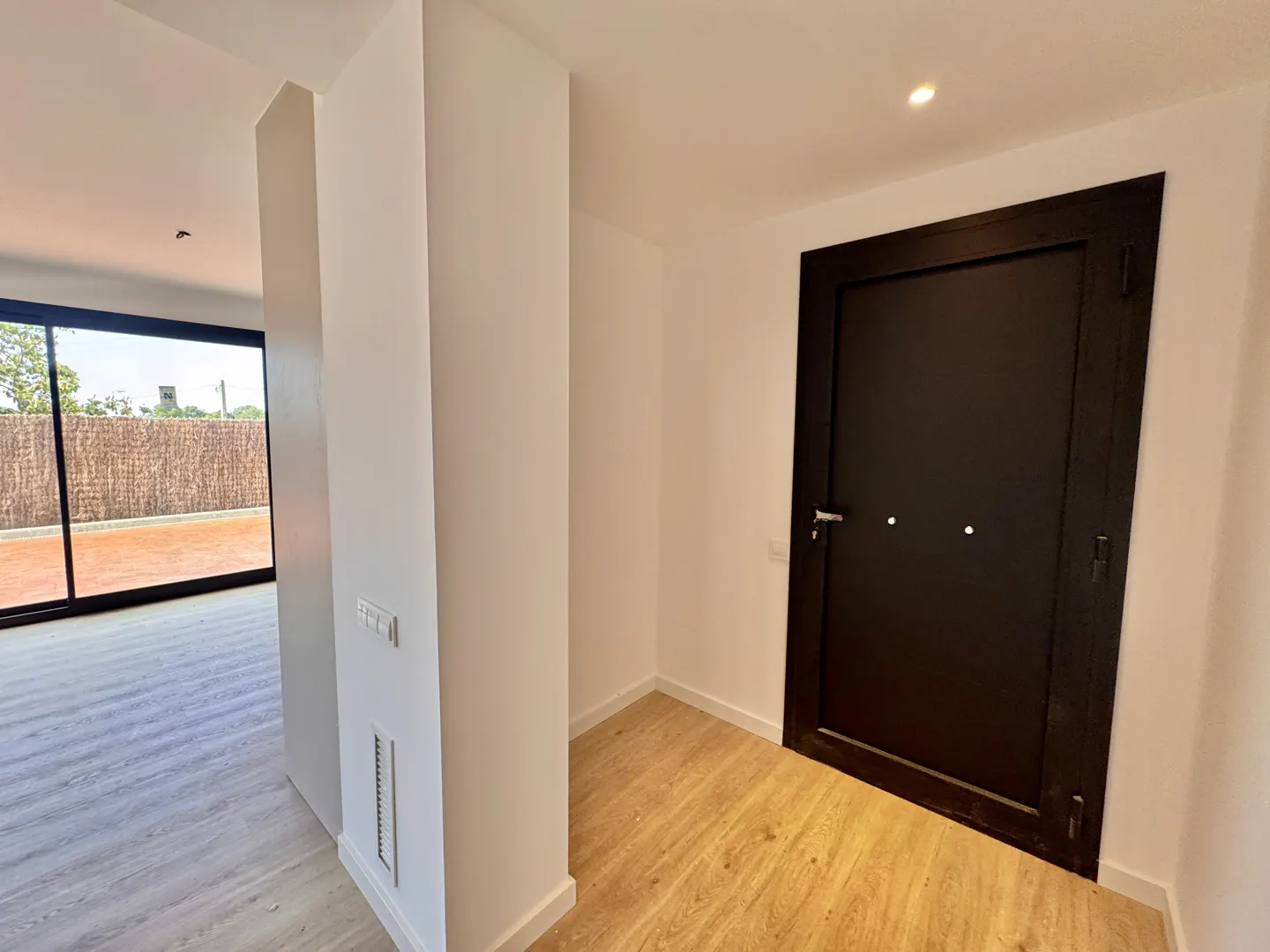 Interior view of a modern home with white walls, light wood floors, and a dark brown door. A large sliding glass door leads to an outdoor patio.