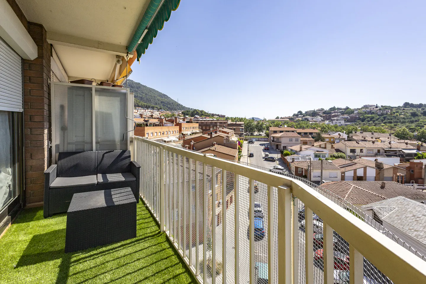 Balcony with artificial grass, black sofa and table. White railing overlooks a town with red-roofed buildings and a green hill in the background.