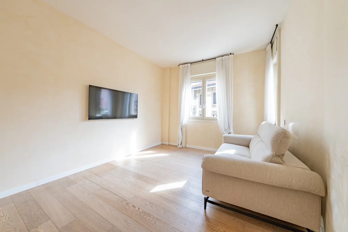 Bright living room with wood floors, cream walls, and a beige sofa. A flat-screen TV hangs on the wall, and a window with white curtains lets in natural light.
