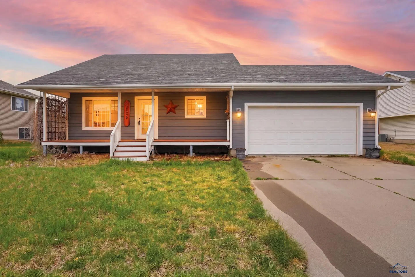 A single-story gray house with a white garage door and porch under a pink and orange sky.