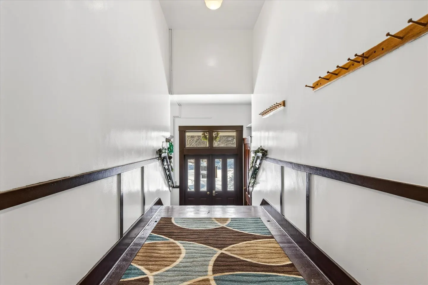 Hallway view with white walls, dark wood trim, and a patterned rug leading to a dark wood door with glass panes. Coat racks are on the wall.