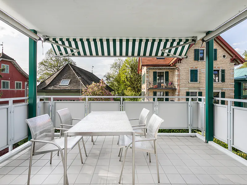 Balcony with white tile floor, metal table and chairs, green and white striped awning, and view of houses.