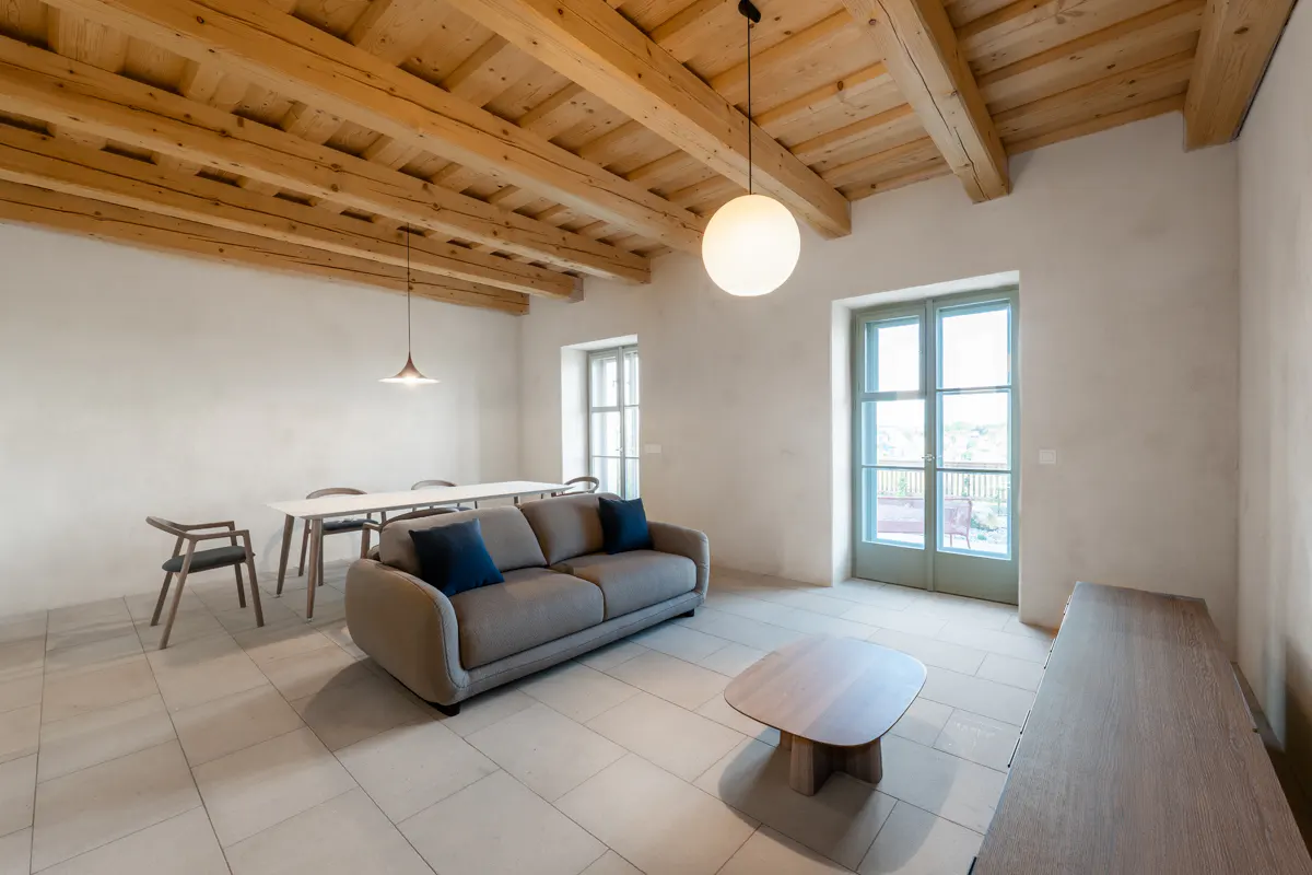 Living room with exposed wood beams, tile floor, and neutral walls. A gray sofa with blue pillows faces a wood coffee table. Dining table and chairs are in the background.