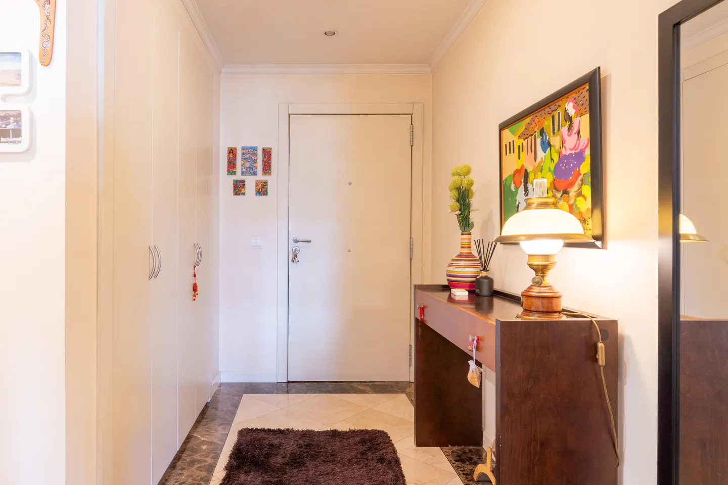 Hallway with white closets, a front door, and a dark wood console table with a lamp, vase, and framed art. A brown rug sits on the marble floor.