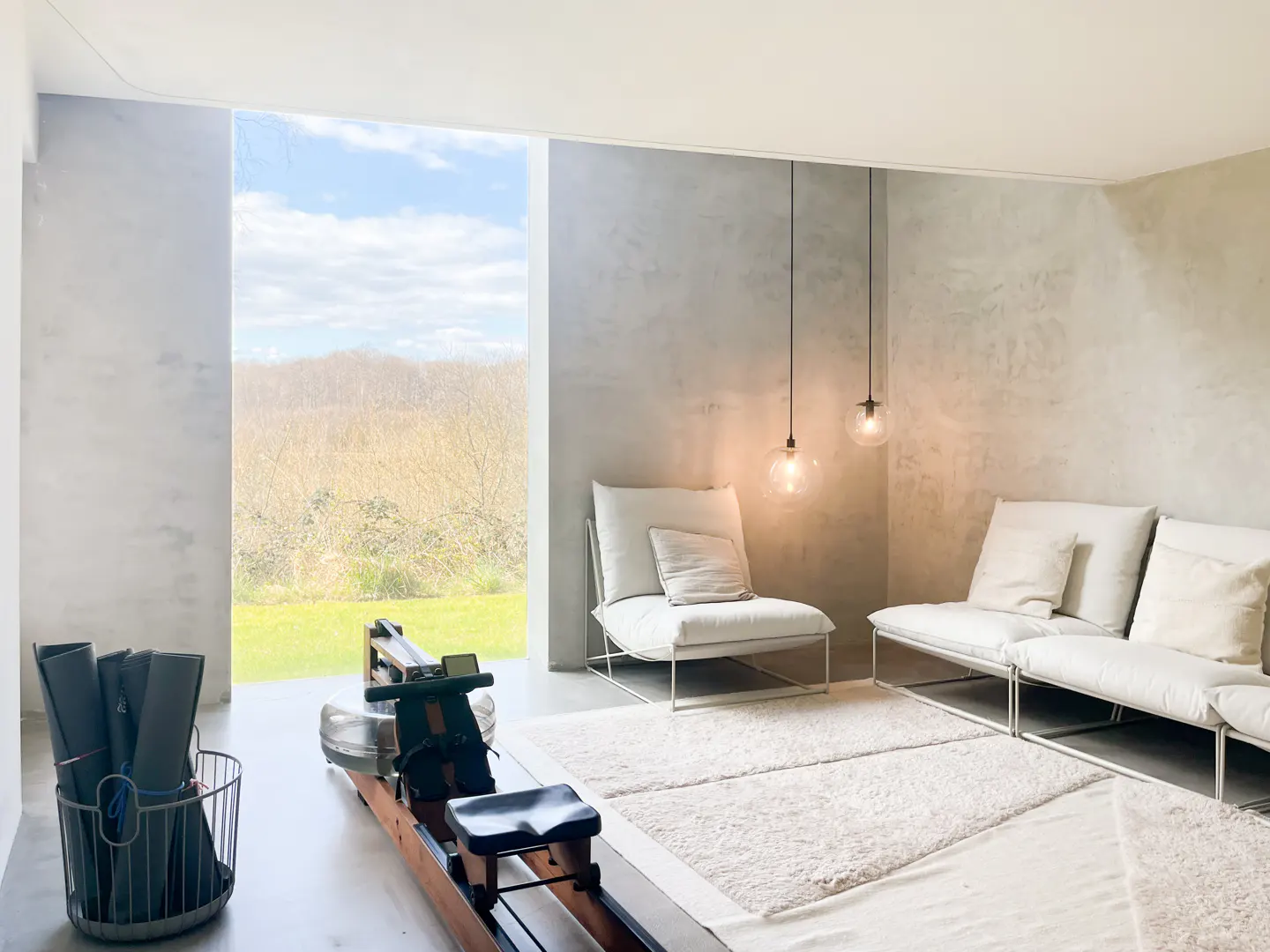 Minimalist room with a rowing machine, white sofa, chair, and a view of a green field through a large window.