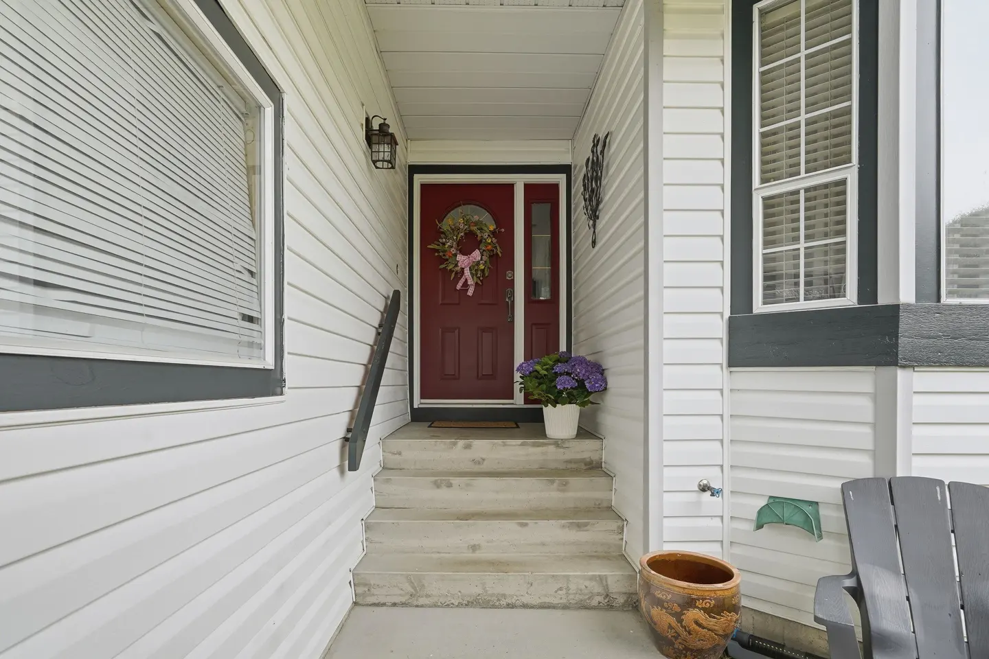 Exterior view of a home's entrance with white siding, a red door with a wreath, and concrete steps. A window with blinds is on the left.