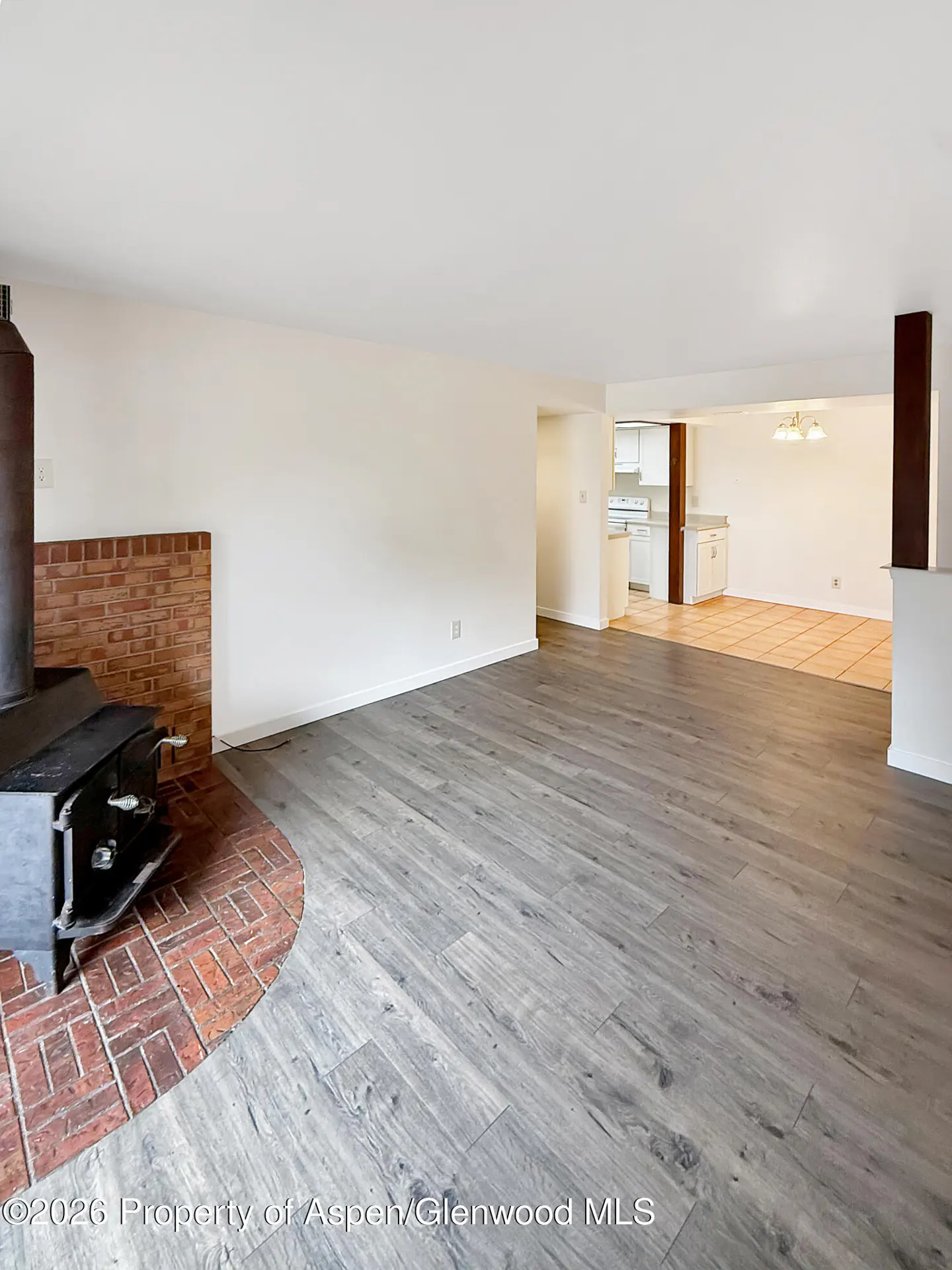 Open-concept living room with gray wood floors, white walls, and a brick fireplace with a black wood-burning stove. Kitchen in background.