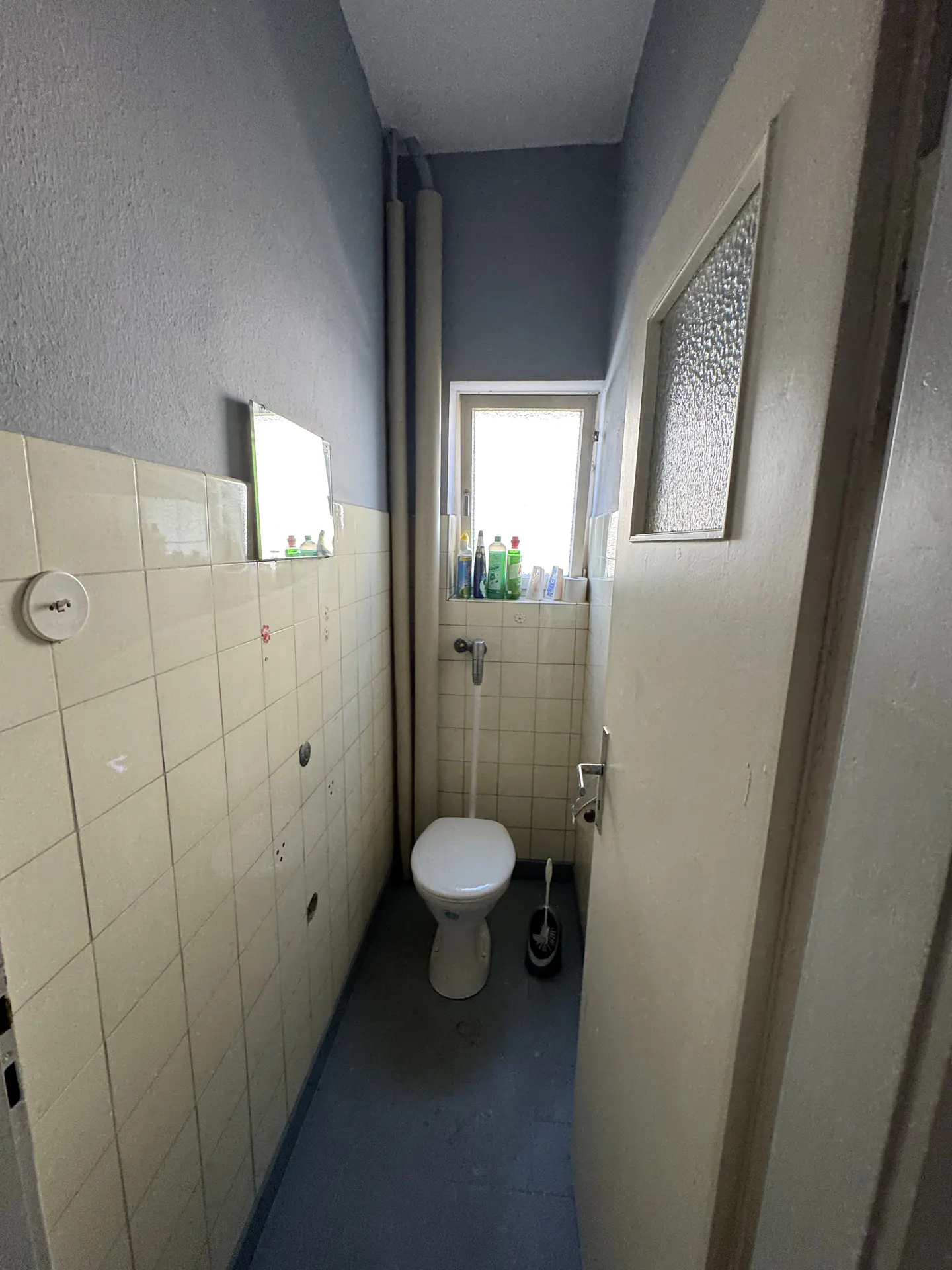 A small bathroom with a white toilet, tiled walls, and a window with cleaning supplies. The walls are gray above the tiles.