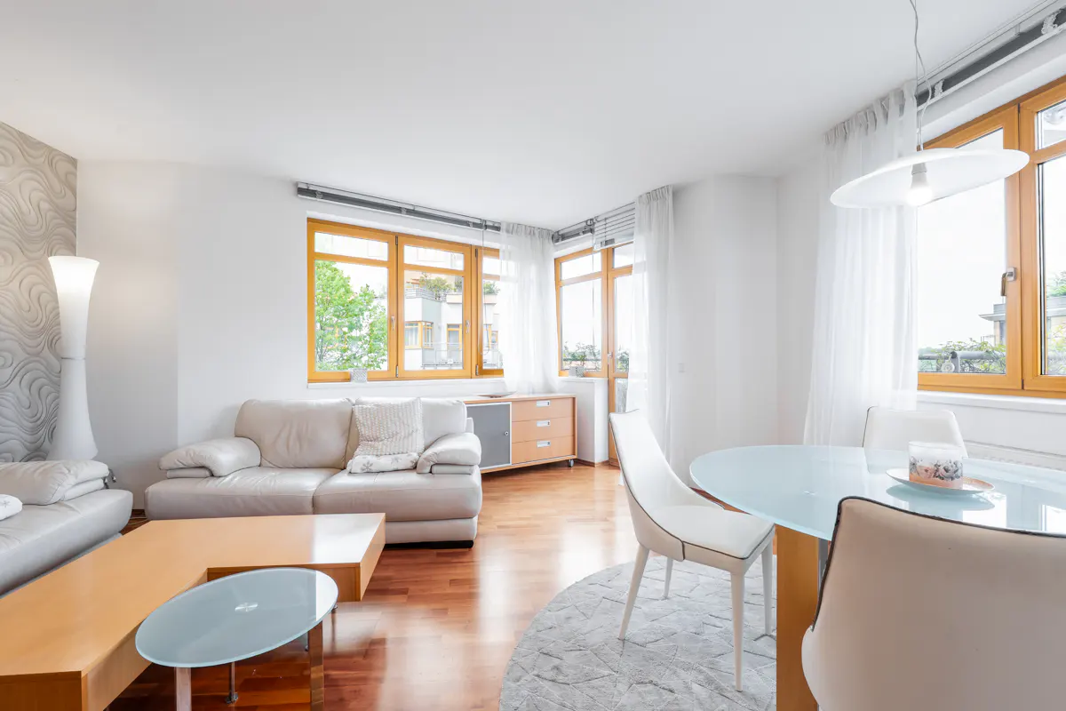 Bright living room with white walls, wood floors, and large windows. Beige sofas, a glass-topped table, and a round dining table with white chairs.