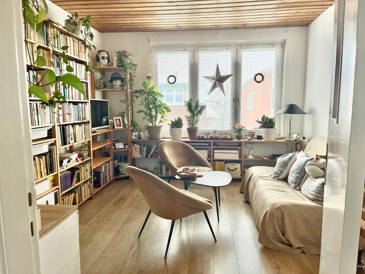 Cozy living room with bookshelves, plants, and two tan chairs around a black table. A beige sofa sits near a window with a star decoration.