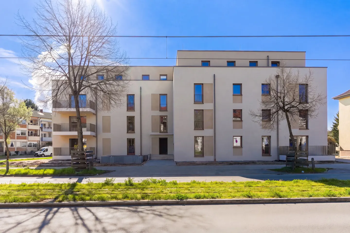 Modern apartment building with beige facade, blue windows, and small balconies under a clear blue sky. Trees frame the building.