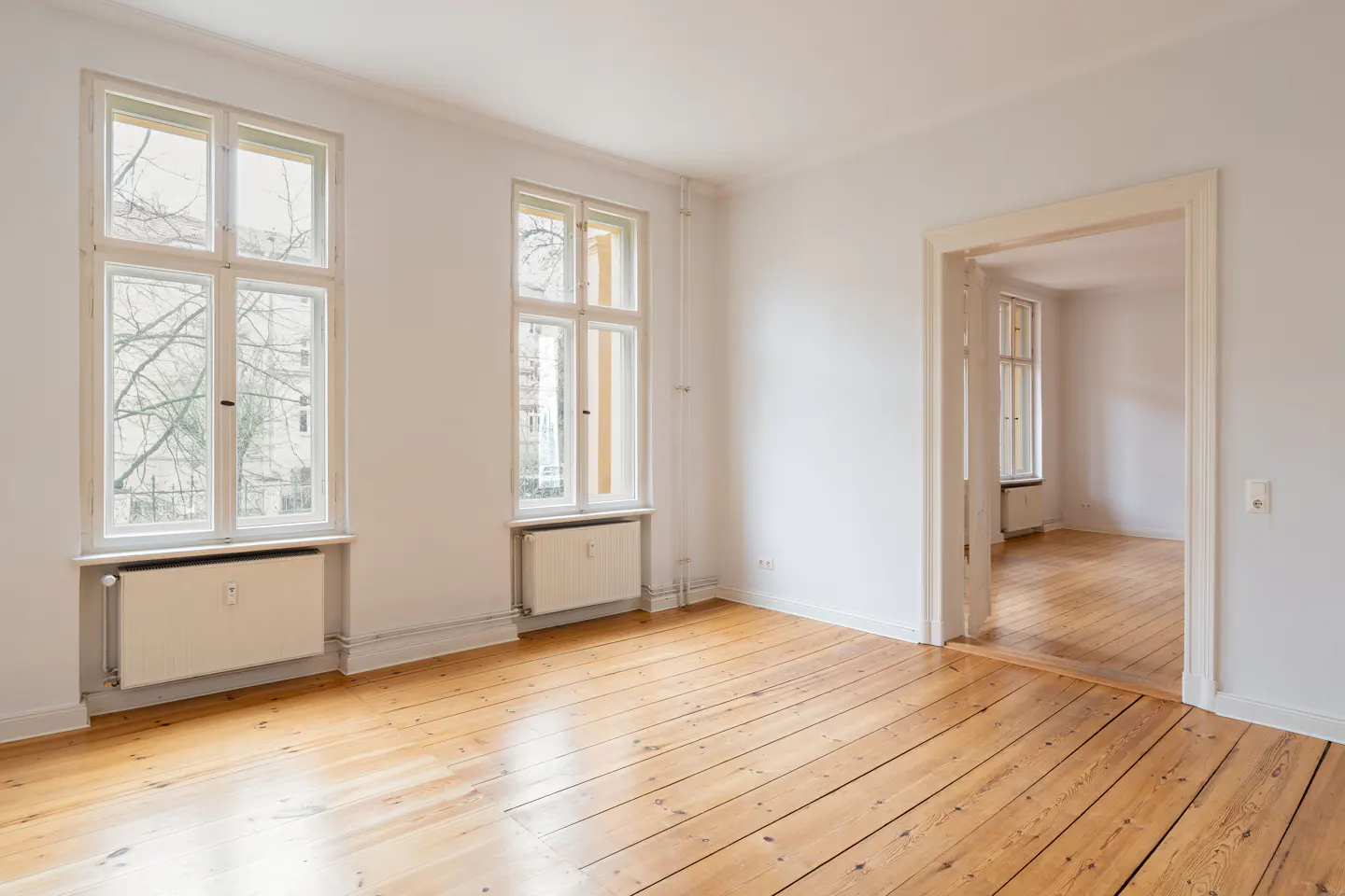 Bright, empty room with hardwood floors, white walls, two windows, and a doorway leading to another room.
