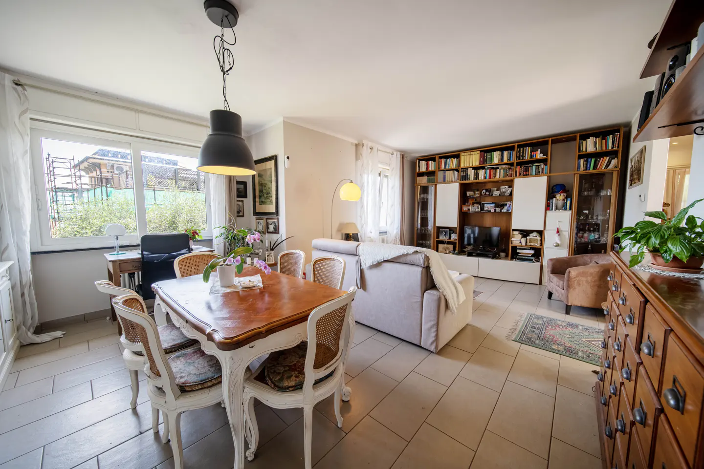 A bright living room with a wooden dining table, white chairs, a beige sofa, and a large bookcase.