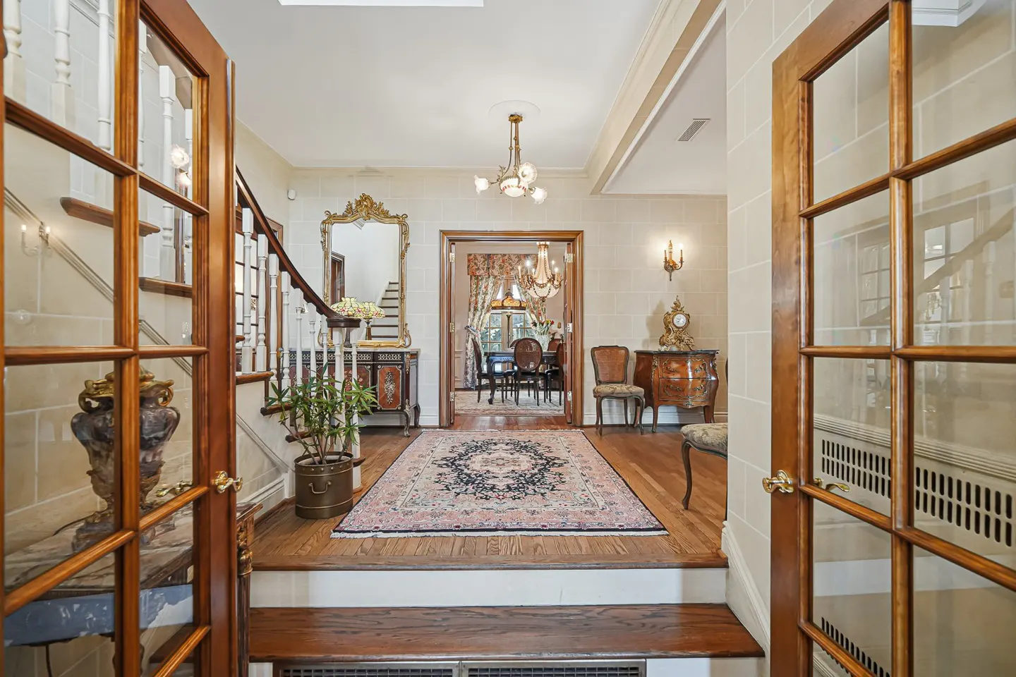 Open wood-framed glass doors reveal a foyer with a staircase, ornate mirror, rug, and dining room beyond.