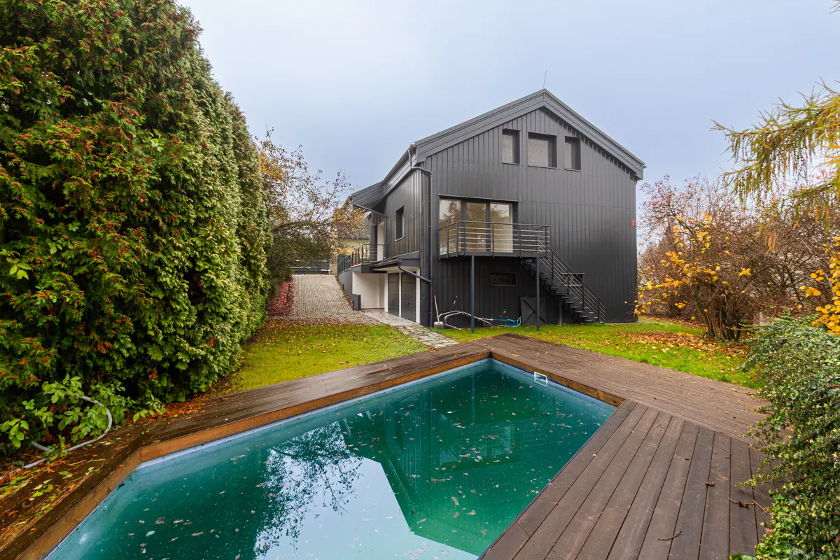 A modern, dark gray house with a pool and wooden deck, surrounded by green trees and autumn foliage.
