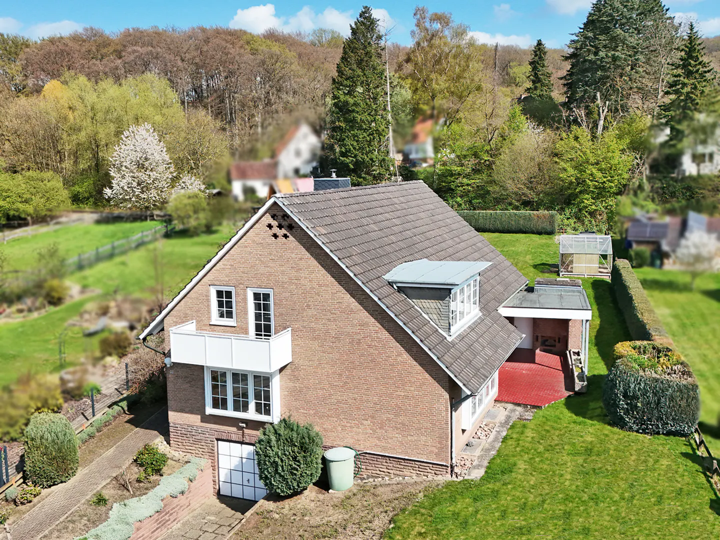 Aerial view of a brick house with a gray roof, white balcony, and green lawn, set against a backdrop of trees and blue sky.