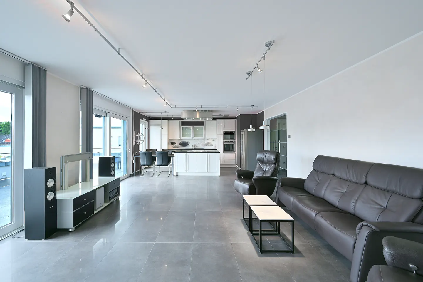 Open-concept living space with gray tile floors, a black leather sofa, and a white kitchen in the background.