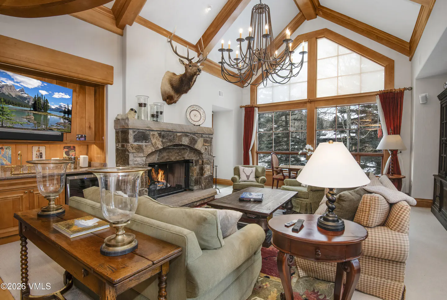 Rustic living room with stone fireplace, elk mount, vaulted wood ceilings, large windows, and comfortable seating.