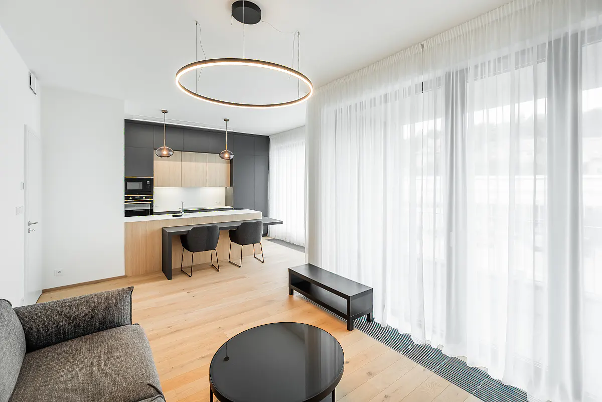 Modern open-plan living area with light wood floors, a sleek kitchen, black bar stools, and a large circular pendant light.