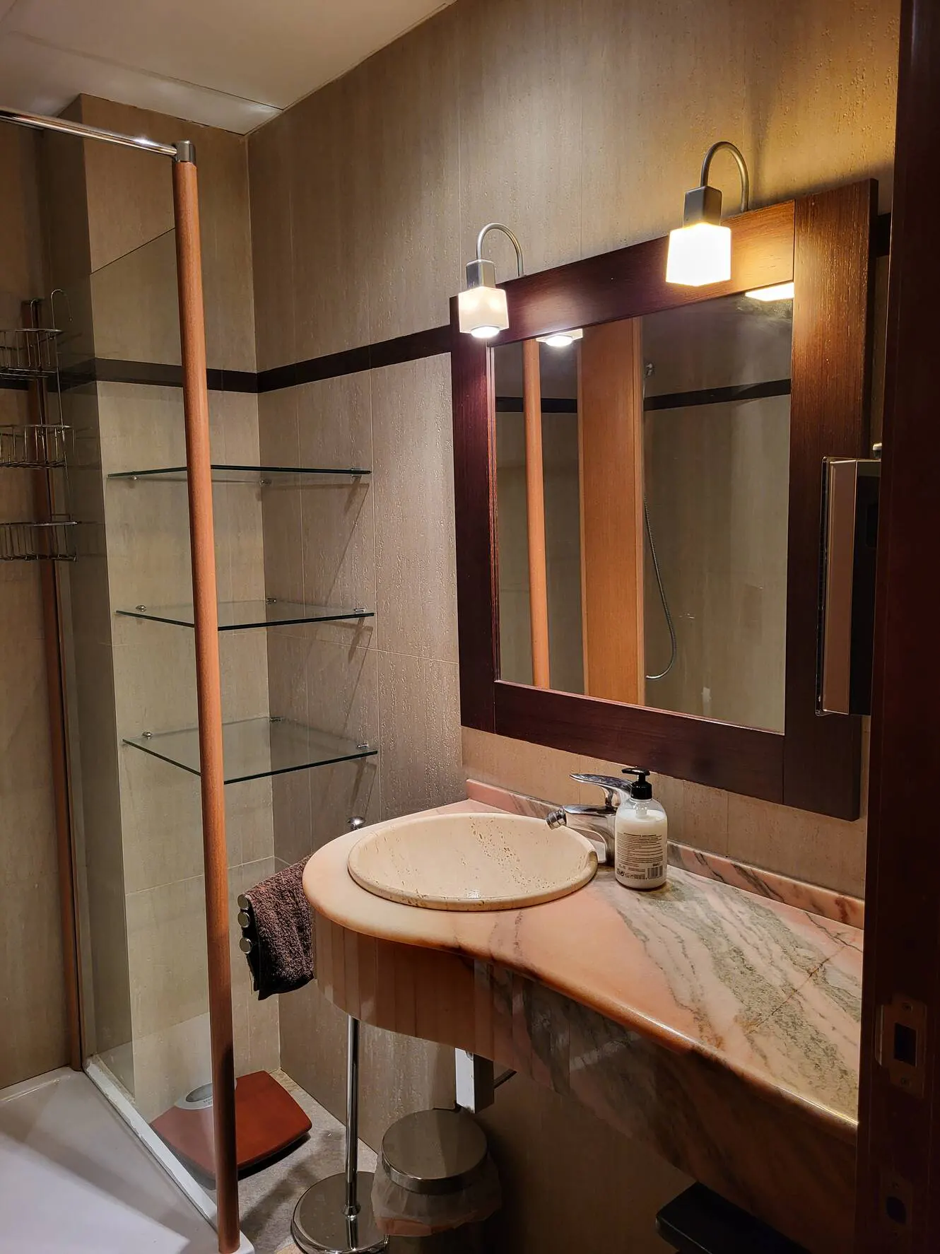 Bathroom with glass shower, marble sink, and dark wood mirror. Shelves are on the wall.