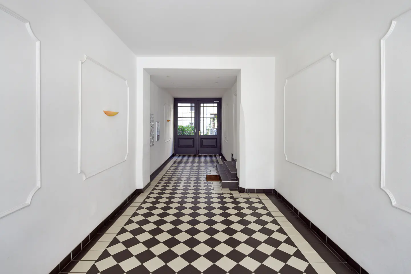 Hallway with black and white checkered tile floor, white walls with decorative molding, and a dark wood door at the end.