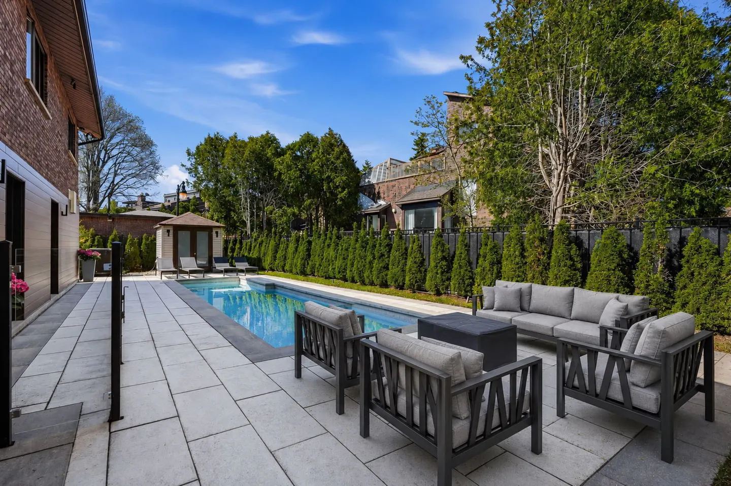 Backyard with a pool, gray patio furniture, and a small cabana, all surrounded by green trees and a black fence.