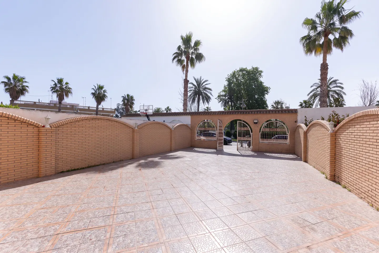 Exterior view of a property featuring a brick wall with arched openings and a tiled courtyard, palm trees in the background.