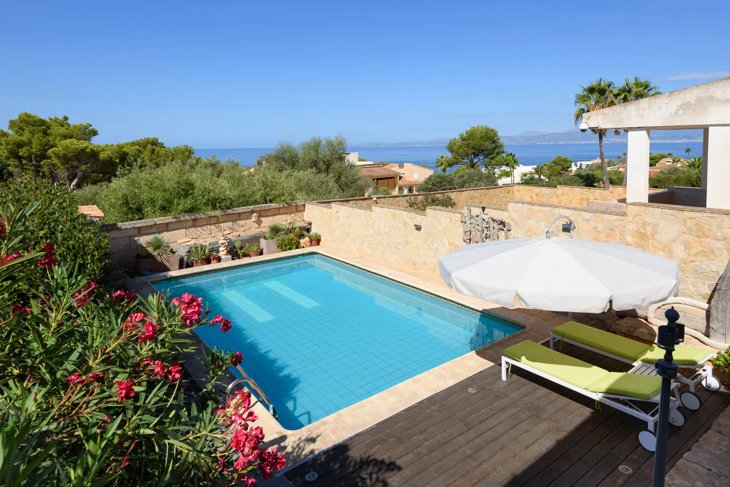 Outdoor pool with blue water, surrounded by stone walls, plants, and lounge chairs under a white umbrella, with ocean view.