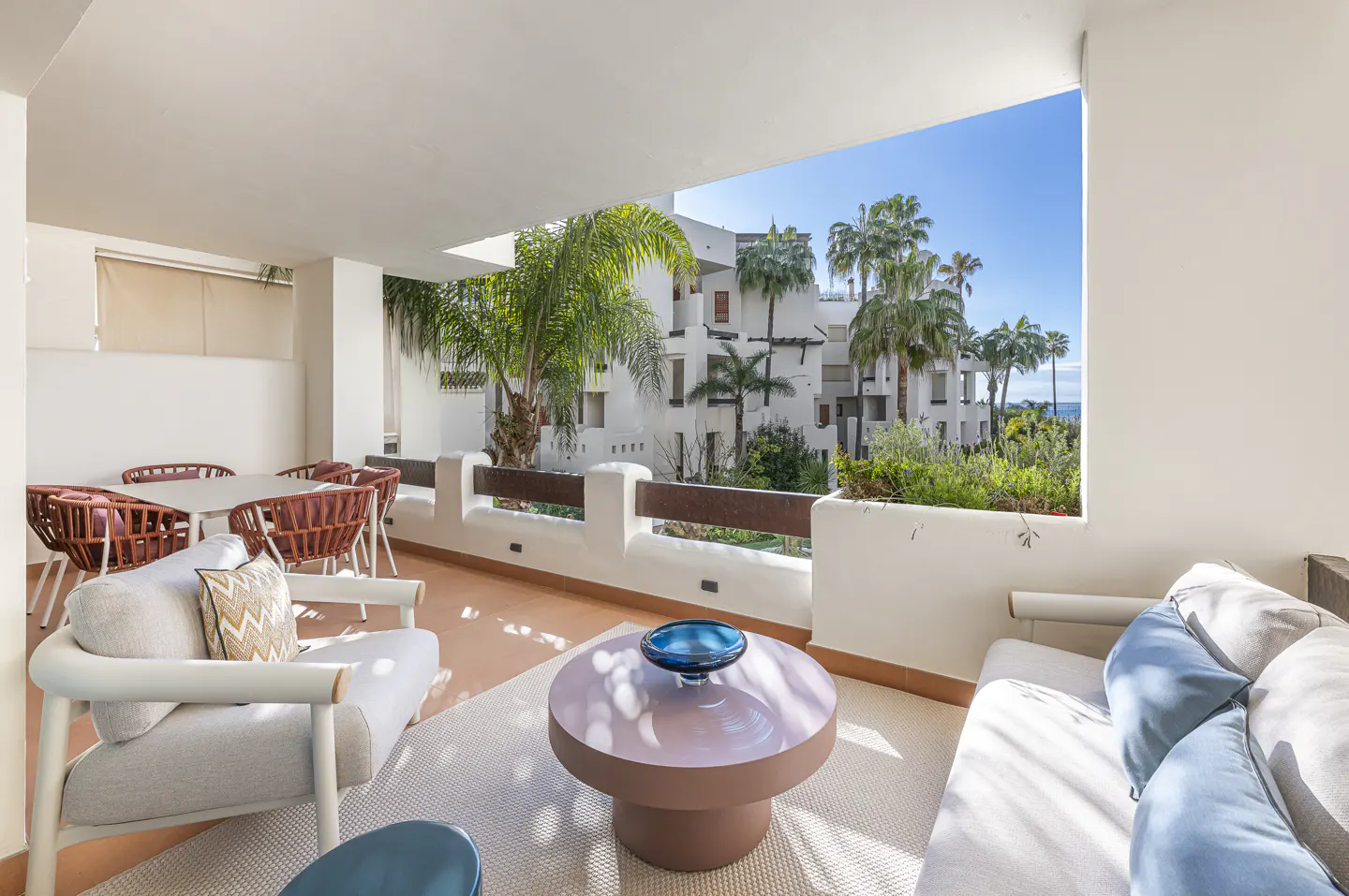 Balcony with outdoor seating, including a table, chairs, sofa, and coffee table. Palm trees and buildings are visible in the background.