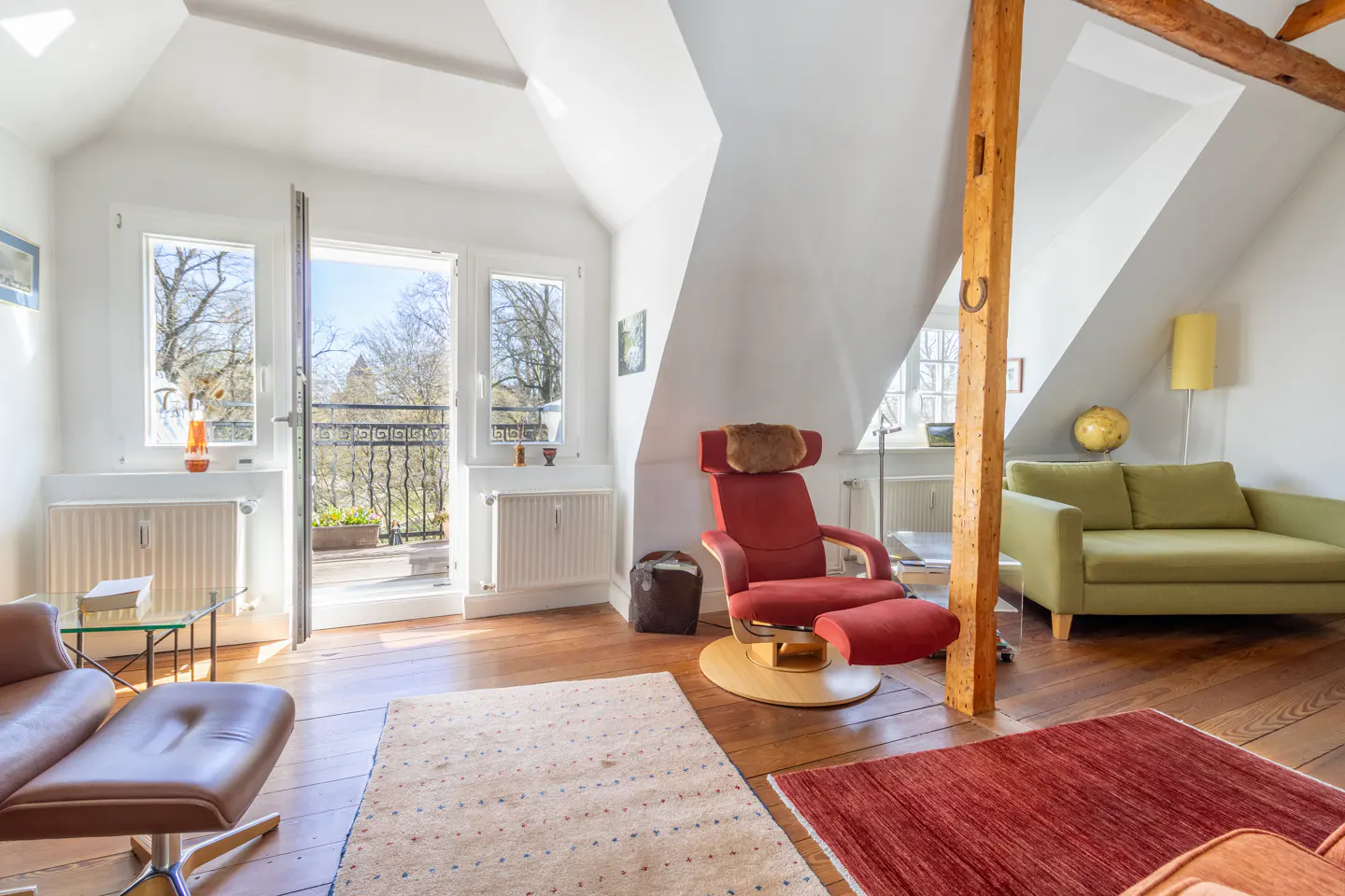 Bright attic living room with white walls, wood floors, and exposed beam. Red chair, green sofa, and open balcony with trees visible.