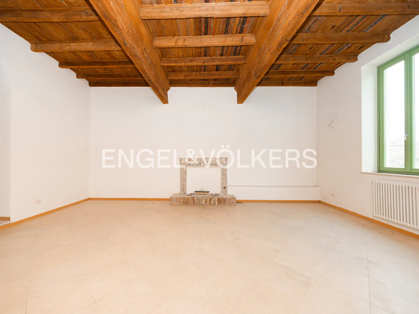 Empty room with white walls, light tan floor, wooden beamed ceiling, stone fireplace, green window frame, and white radiator.