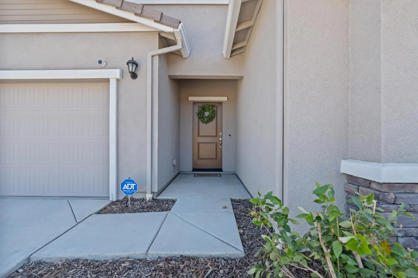 Beige house exterior with a brown front door with a wreath, a gray garage door, and an ADT security sign.