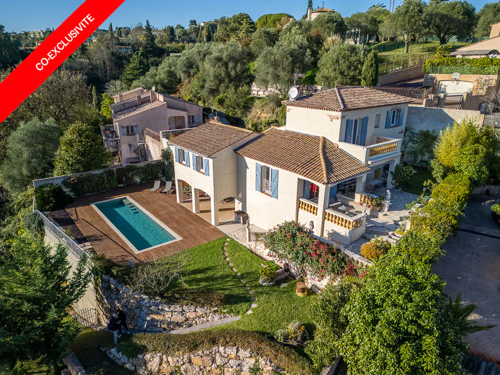 Aerial view of a white house with blue shutters, a brown tile roof, and a pool surrounded by trees. "Co-Exclusivite" banner.