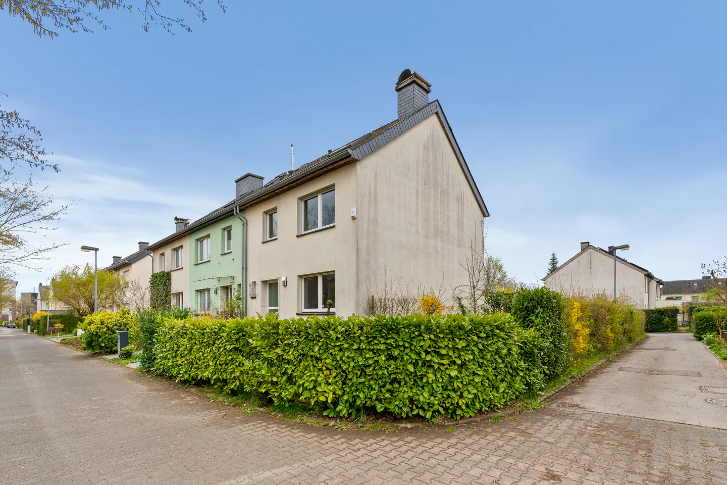 Exterior view of a beige and light green row house with a tall green hedge in front, set on a paved street under blue sky.