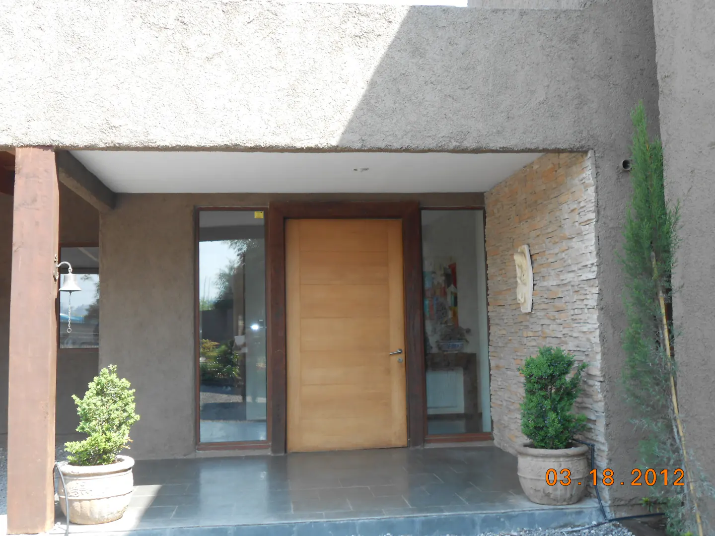 Front entrance of a modern home with a light wood door, stone accent wall, and potted plants.