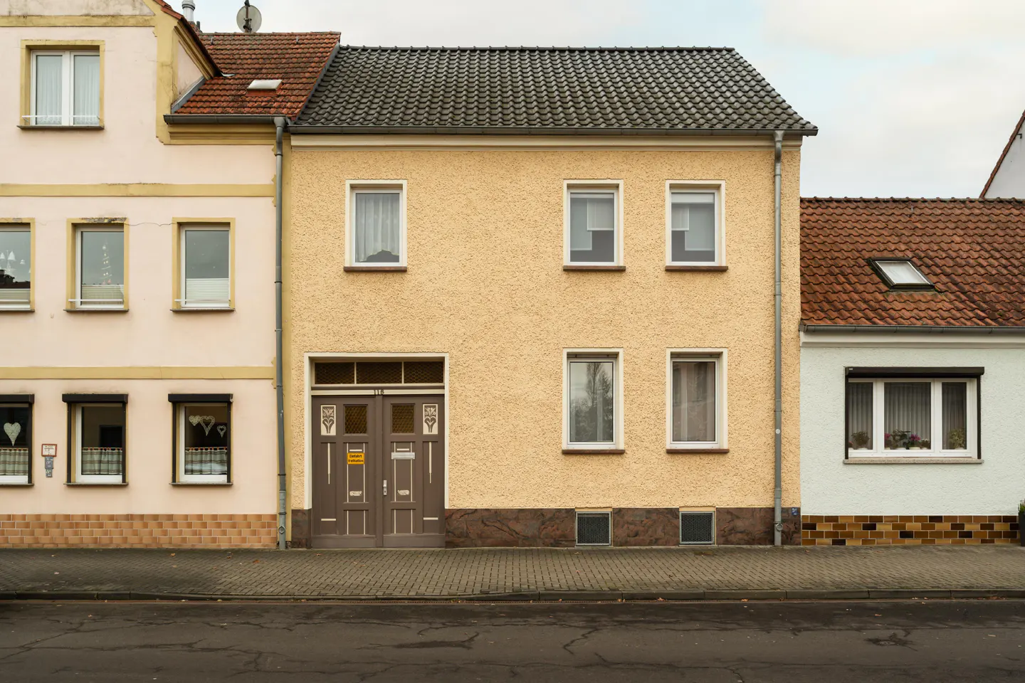 A two-story yellow house with a gray roof and brown double doors, flanked by other houses.