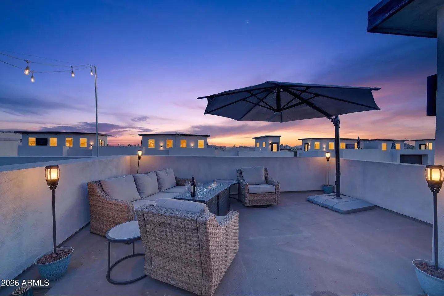 Rooftop patio at dusk with wicker furniture, fire table, and umbrella. String lights and tiki torches add ambiance. Buildings visible in the background.