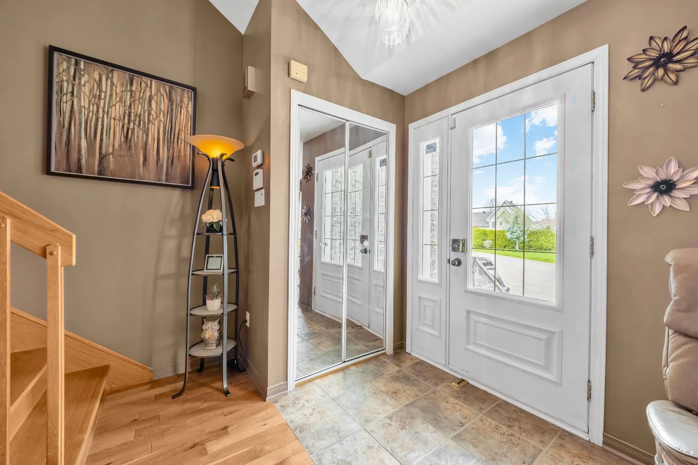 Foyer with tan walls, tile floor, and a white front door with glass panes. A mirror closet door reflects the interior. A floor lamp and stairs are on the left.