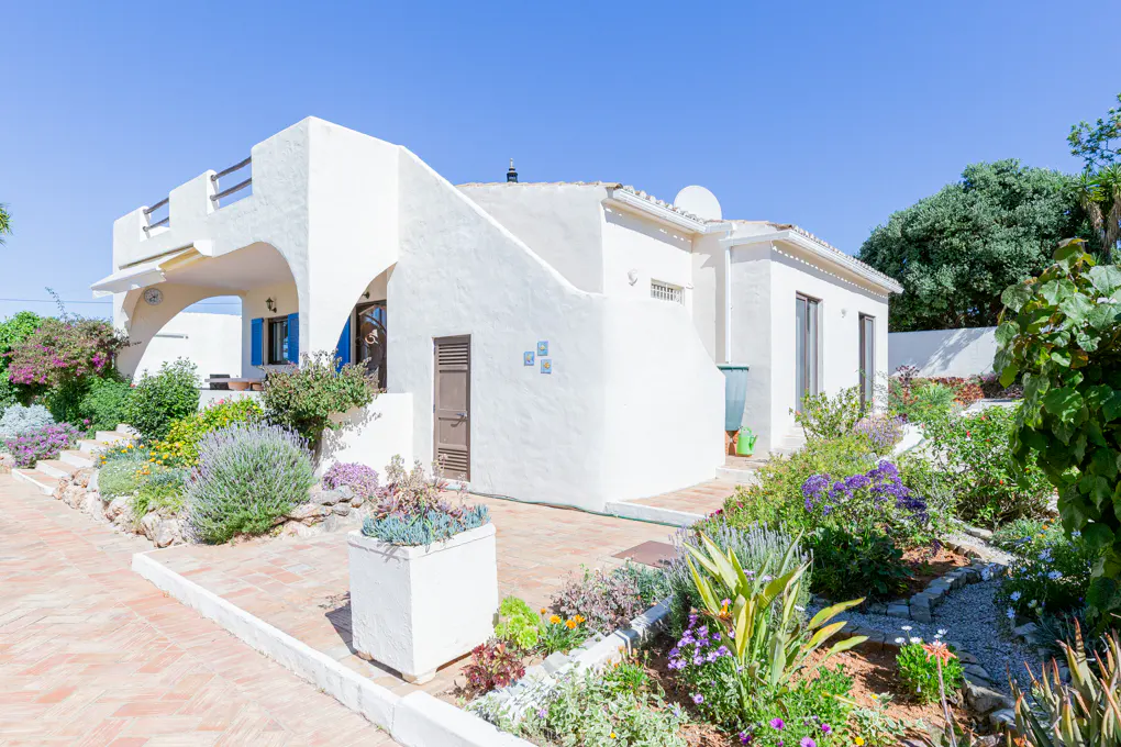 Exterior of a white stucco house with a red brick walkway and lush garden under a clear blue sky.