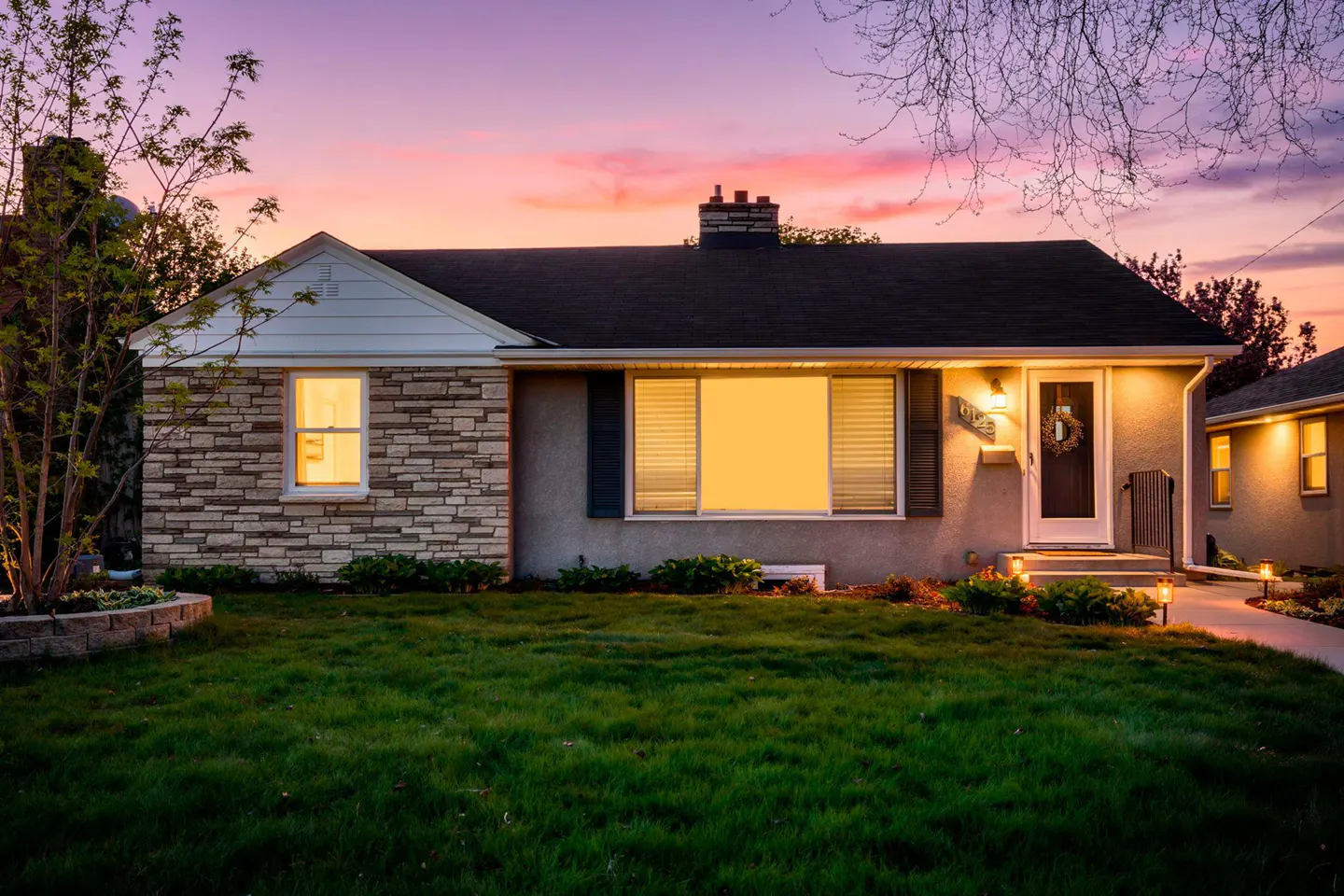 A ranch-style house with stone and stucco exterior at dusk, lights on, and a green lawn.
