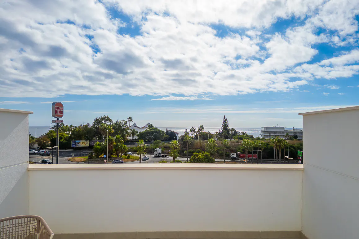 View from a white balcony overlooking a road with cars, trees, and the ocean under a blue sky with white clouds. A Burger King sign is visible.