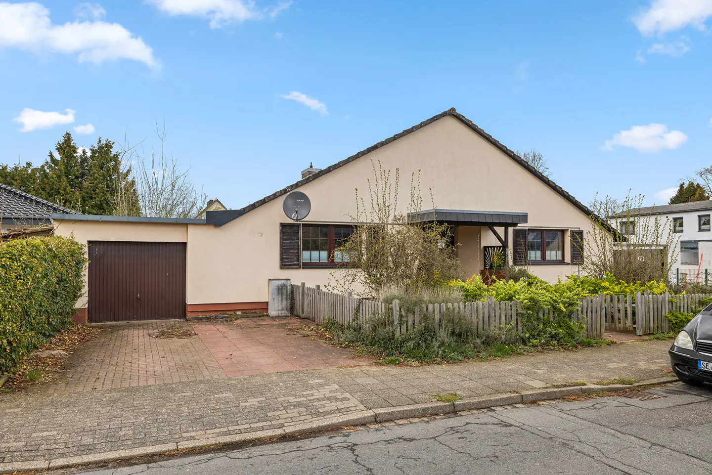 Beige single-story house with a brown garage door, a satellite dish, and a small wooden fence.