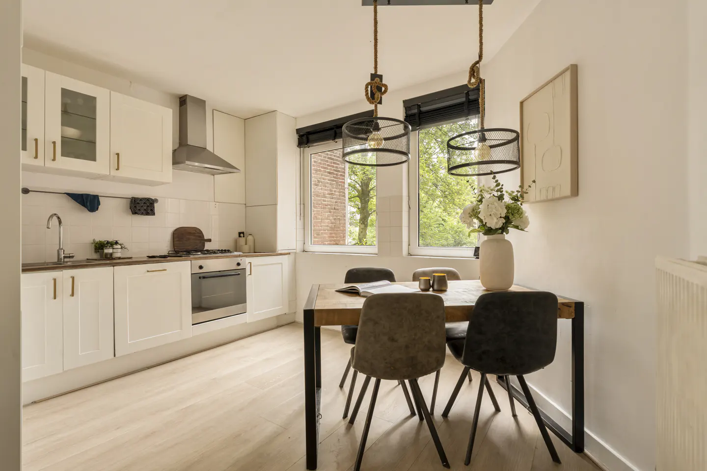 Bright kitchen with white cabinets, stainless steel appliances, and a wooden dining table with four chairs under two hanging lights. A vase of white flowers sits on the table.