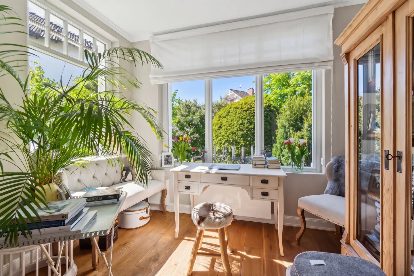 Bright sunroom office with white desk, stool, and bench. Large windows overlook a lush green garden. A tall palm adds a tropical touch.
