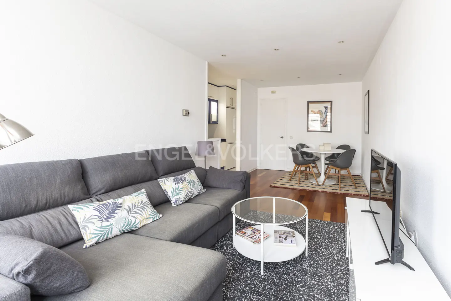 Living room with a gray sectional sofa, a round glass coffee table, and a dining table with four chairs. The walls are white, and the floor is wood.
