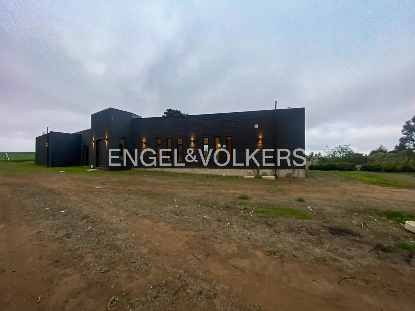 Modern, dark-colored home with Engel & Volkers logo on the front. The sky is cloudy and the ground is dirt.