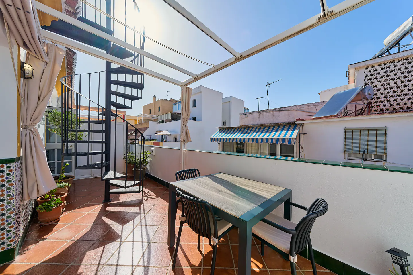 Outdoor patio with a table, chairs, and spiral staircase. The patio has terracotta tiles and a white wall. Buildings are visible in the background.